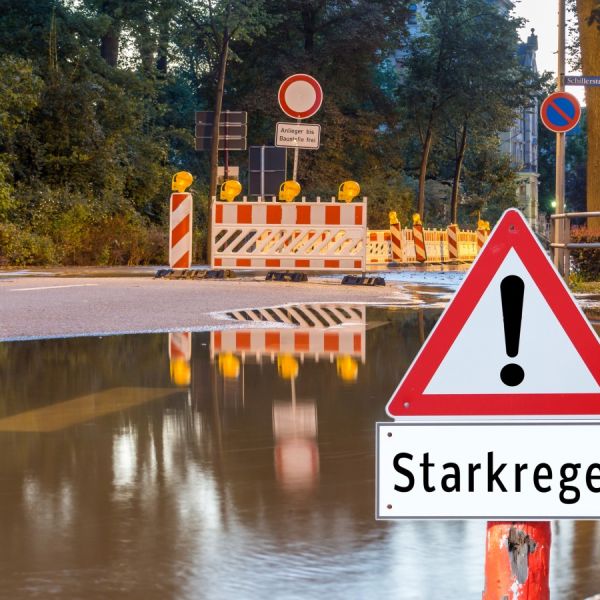 Überflutung u.a. einer Schillerstraße durch Starkregen. Achtung Starkregen Warnschild! Baustellenschild mit "Anlieger bis Baustelle frei" Einschränkungshinweisschild, umgeben von Absperrzäunen und -baken z.T. mit Warnbeleuchtung in einer Stadt.